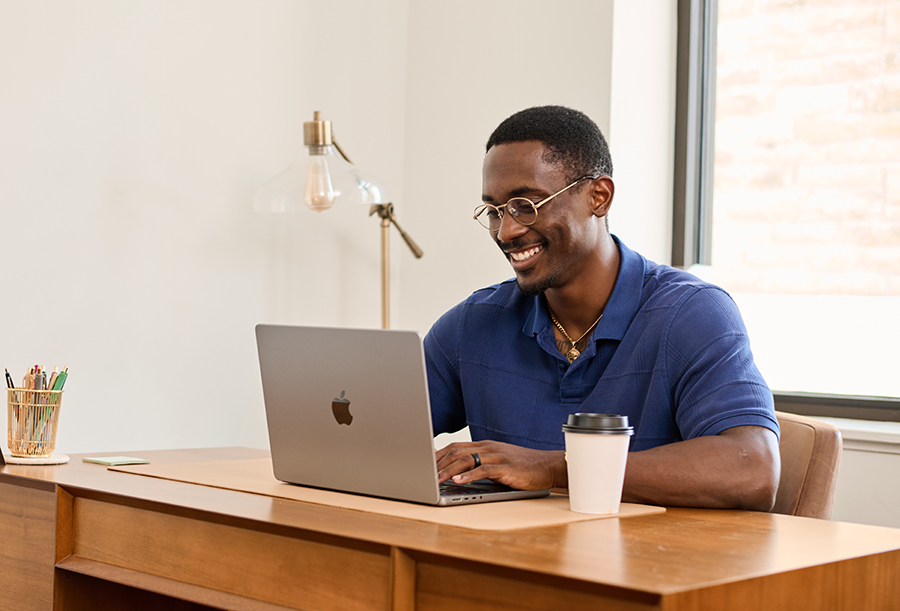 A man in a blue shirt is typing on a laptop. He is sitting at a desk and has a cup of coffee next to him.