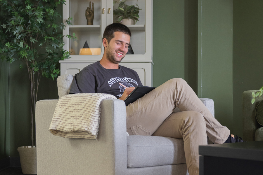 A man is sitting in a chair working on a tablet. He is smiling and wearing a gray SEU shirt.