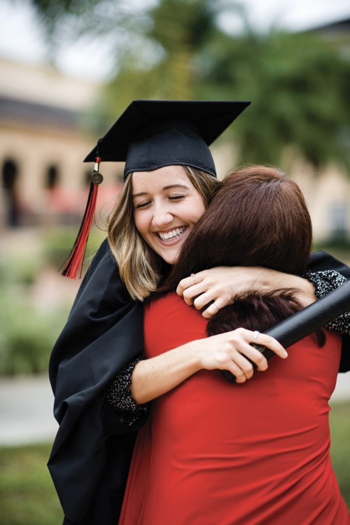 Graduate smiling wearing seu graduation attire in black and red hugging mom in red SEU shirt