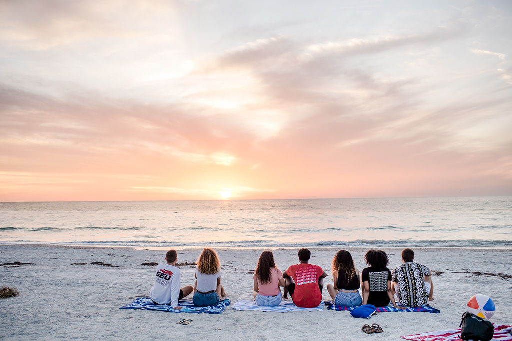 Students sitting on the sand at a Florida beach during sunset.