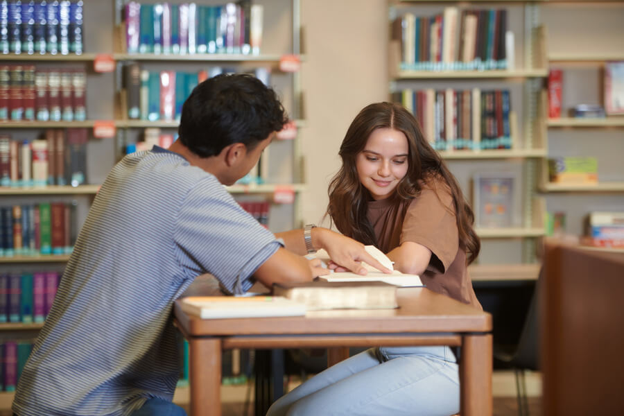 Two students are sitting in a library looking at a book together. The young man is pointing to a page of the book and the young woman is looking at the page.