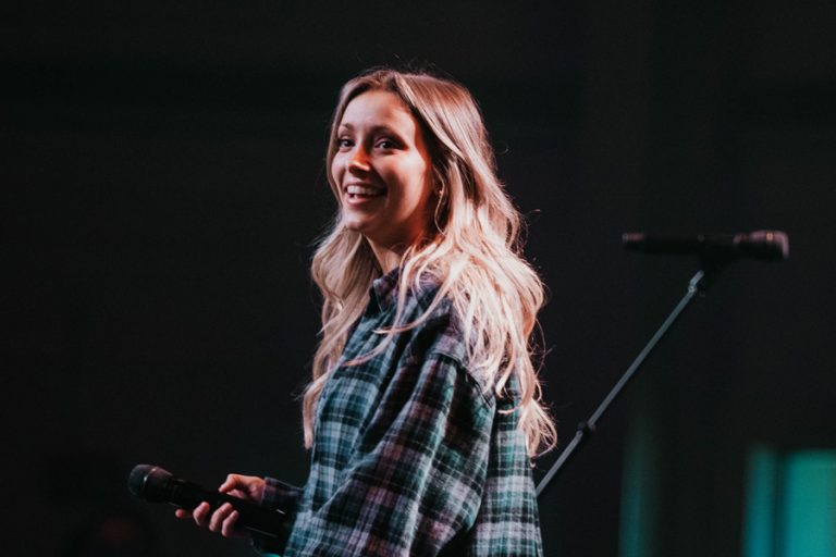 Chelsea Plank standing on stage holding a microphone, wearing plaid shirt.