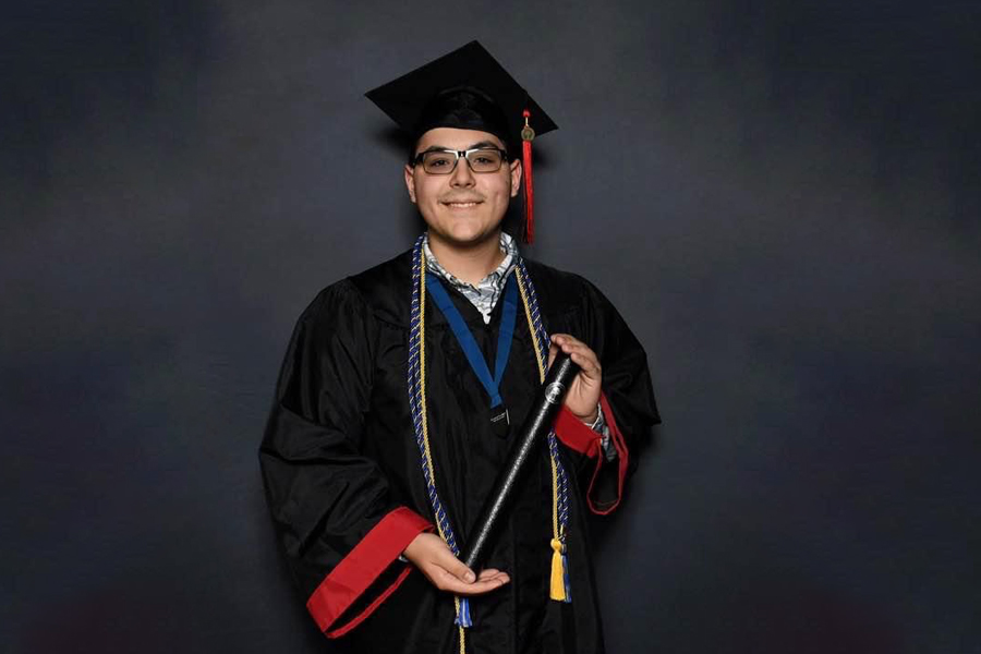 Man in cap and gown holding a diploma in front of a dark grey backdrop.