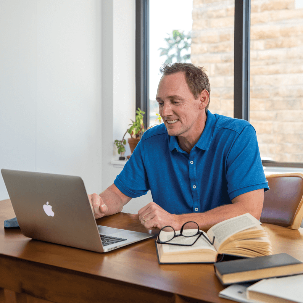 A man is sitting at a desk working on a laptop computer. He is wearing a blue shirt. Open books and a pair of glasses are on the desk next to him