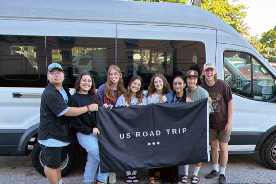 Students smiling in front of white van holding US ROAD TRIP flag