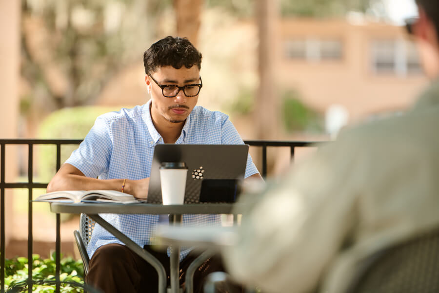Man sitting at a table outside at SEU, working on his computer and drinking his coffee.