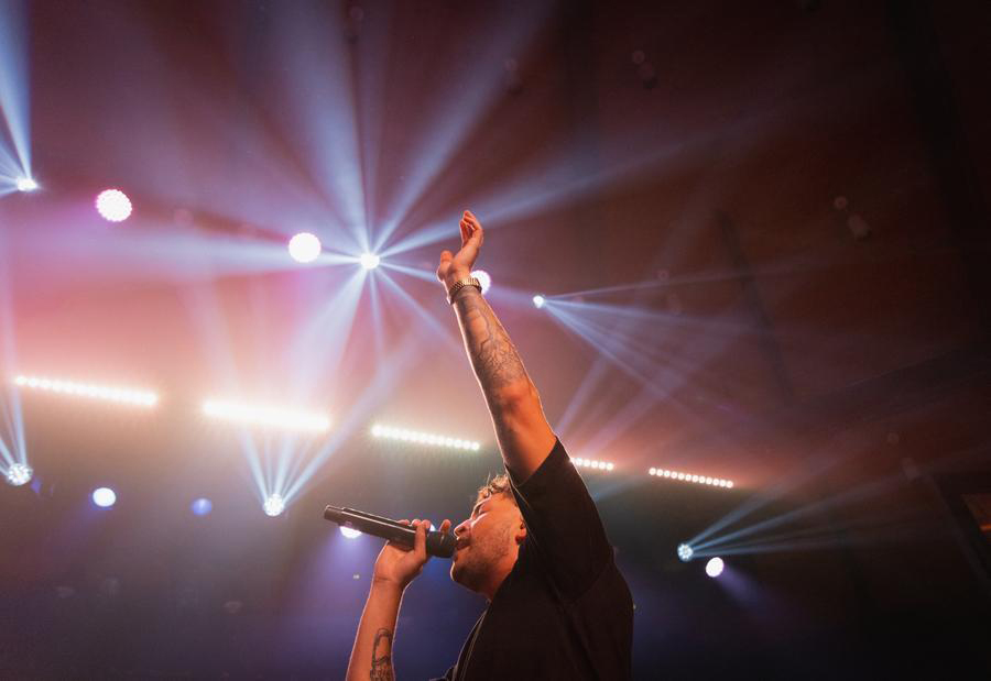 A man in a black shirt is singing into a microphone with his arm raised. Stage lights are above him.