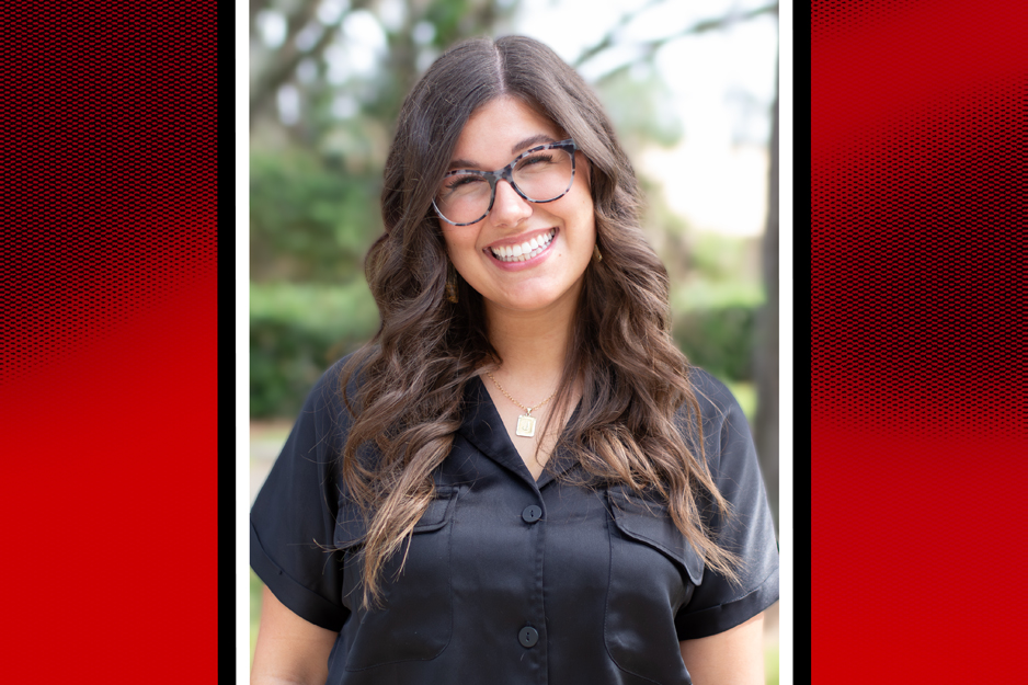 A photo of Jade Beesley smiling. She is wearing a black shirt and glasses and she has curly brown hair.