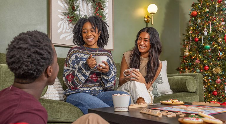 Two young women and a young man are gathered around a coffee table drinking hot chocolate. A Christmas tree is behind the, and a Scrabble board is on the table.