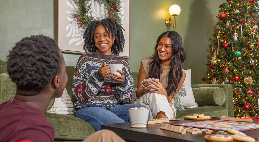 Two young women and a young man are gathered around a coffee table drinking hot chocolate. A Christmas tree is behind the, and a Scrabble board is on the table.