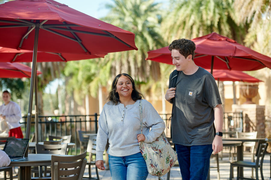 Two students, one female and one male, are walking together in front of some tables with red umbrellas.